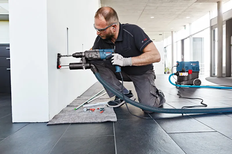 一名佩戴安全设备的人员正在用除尘装置在墙上钻孔。.