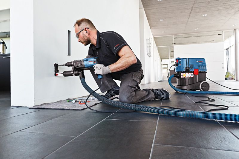 一名佩戴安全设备的人员使用带有除尘功能的电动工具在墙上钻孔。.