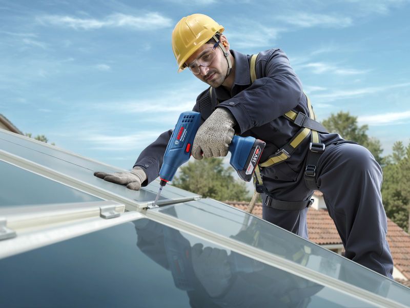 一名佩戴安全设备的人员使用电动工具拧紧屋顶太阳能电池板上的螺栓。.