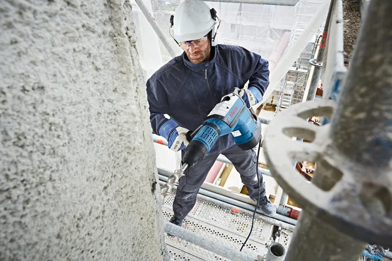 一名佩戴安全设备的人员在脚手架上用旋转锤钻入混凝土。.
