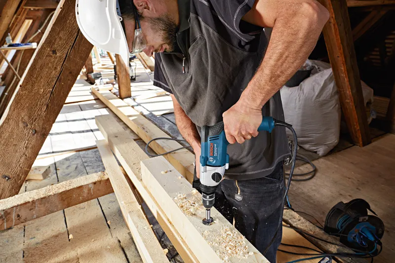 一名佩戴安全设备的人员在木梁上钻了一个大孔。.