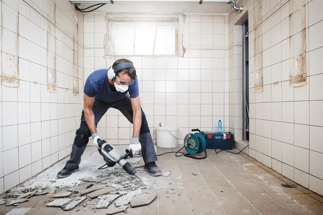 一名佩戴安全设备的工人使用电钻打碎地砖。.