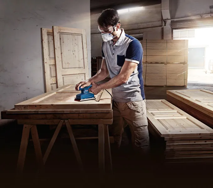 一名佩戴安全设备的人员在车间使用轨道砂光机打磨木门。