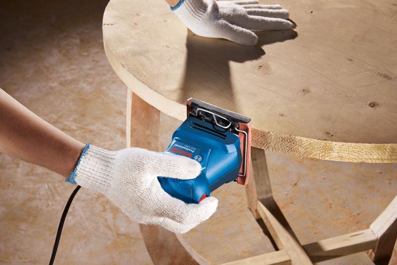 一名佩戴安全设备的人员正在使用轨道砂光机打磨木桌的边缘。