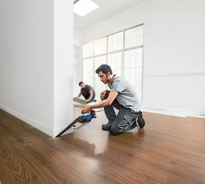 一个人用无绳锯在墙边切割地板，另一个人则安装地板镶板。.