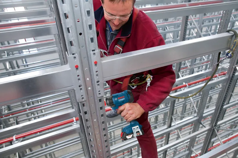 一名佩戴安全设备的人员在仓库中使用电钻钻削金属梁。.