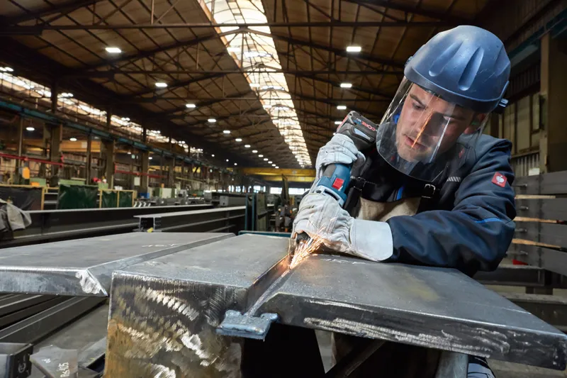 一名佩戴安全设备的工人在工厂里用电动工具研磨金属。.