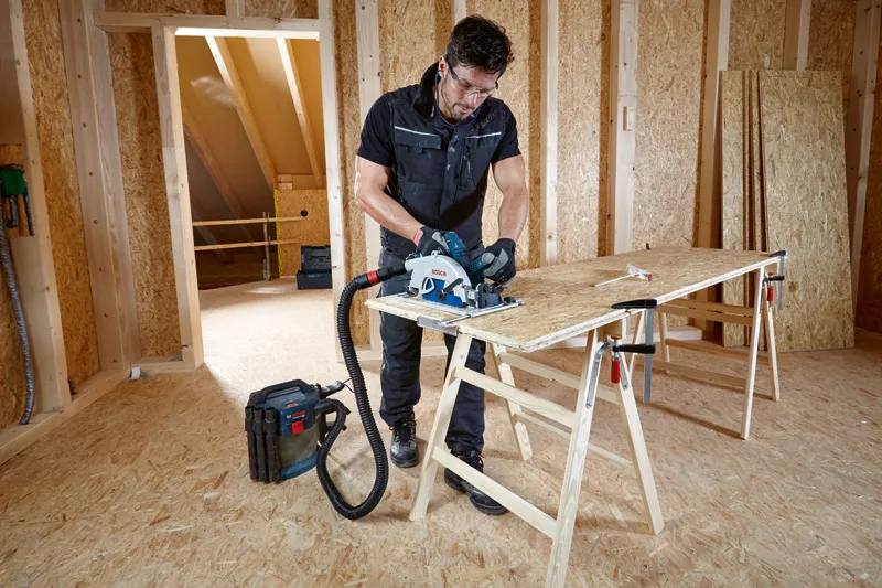 一名佩戴安全设备的人员使用连接到除尘器的圆锯切割胶合板。.