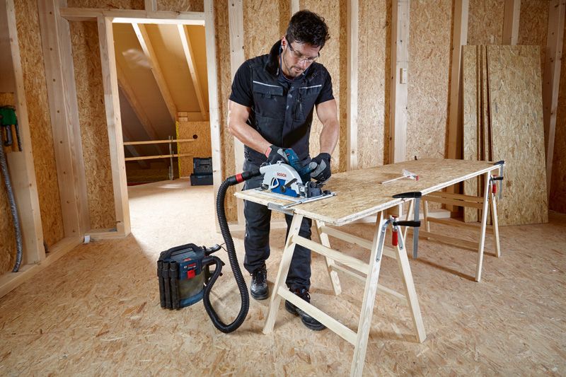 一名佩戴安全设备的人员使用连接到除尘器的圆锯切割胶合板。.