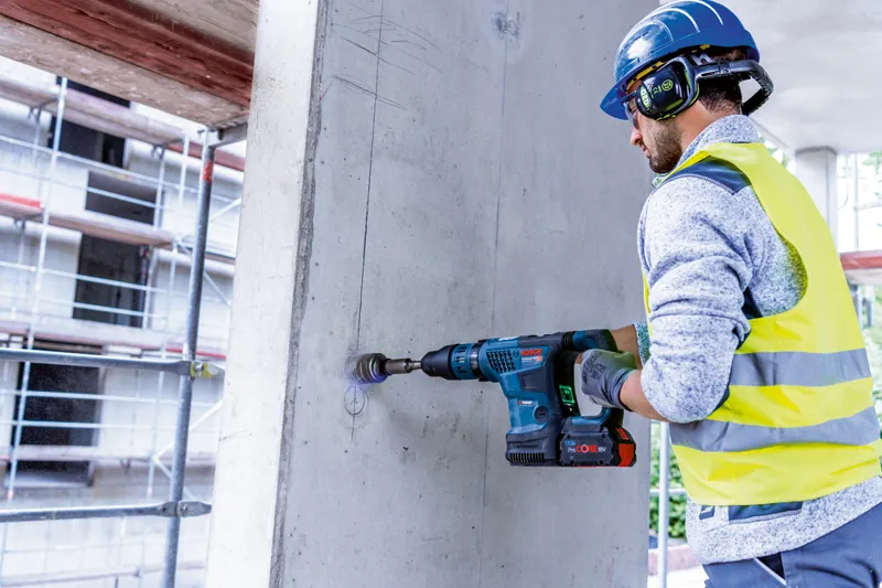 佩戴安全设备的人员使用无绳旋转锤在混凝土墙上钻孔。.