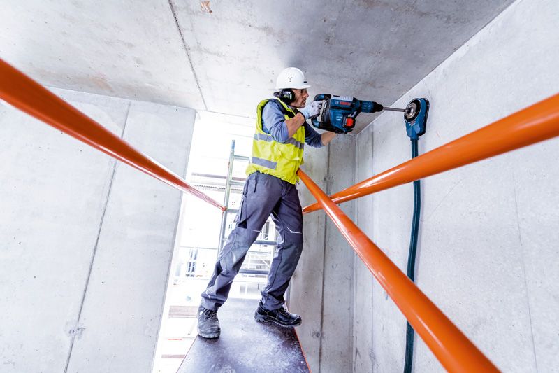 一名佩戴安全设备的人员使用旋转锤在混凝土天花板上钻孔。.