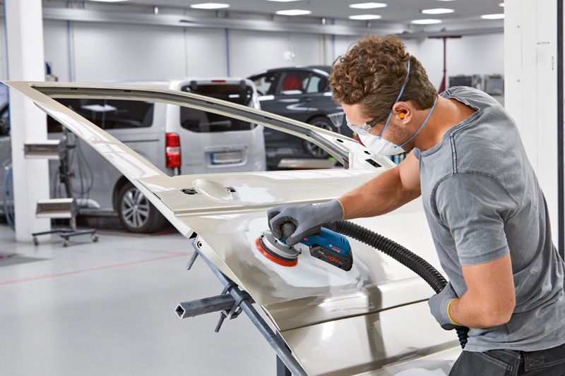 汽车修理厂里，一名佩戴安全设备的人员正在用电动砂光机打磨车门。.