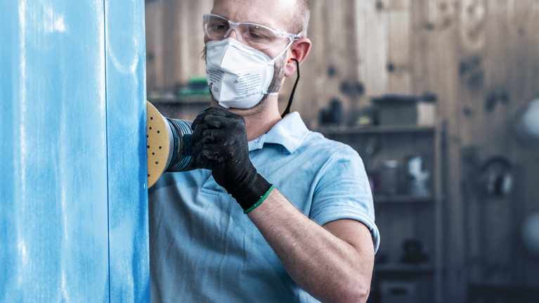 佩戴安全设备的人员使用电动砂光机打磨蓝色面板。