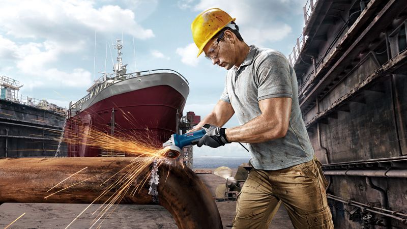 一名佩戴安全设备的人员在造船厂研磨金属管道，产生火花。.