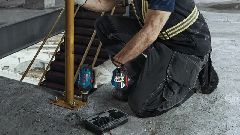 一名佩戴安全设备的人员使用无绳冲击扳手拧紧金属柱上的螺栓。.