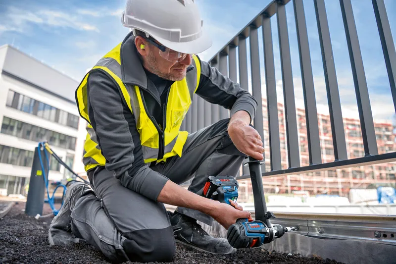 一名佩戴安全设备的人员使用无绳组合钻在金属栅栏框架上钻孔。.