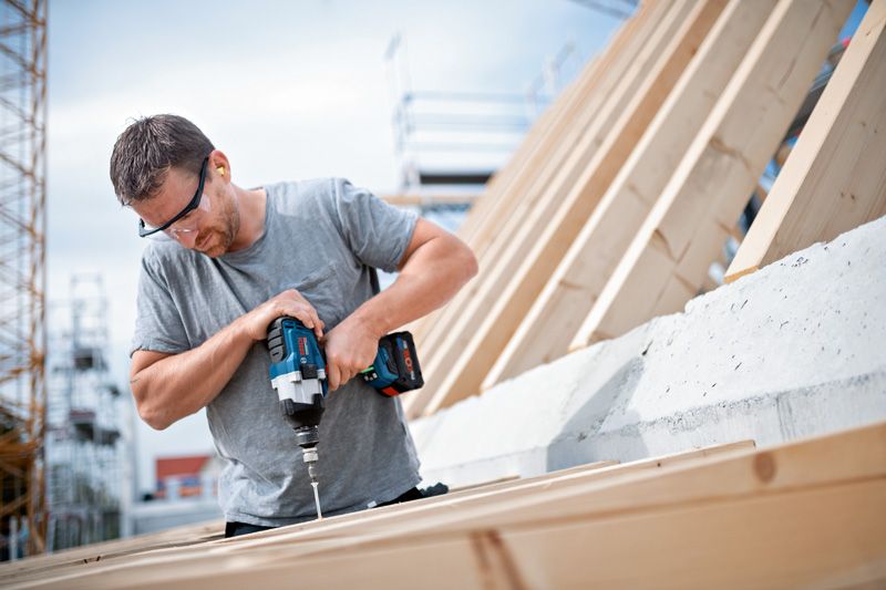 佩戴安全设备的人员使用无绳冲击扳手将螺丝拧入木头。.