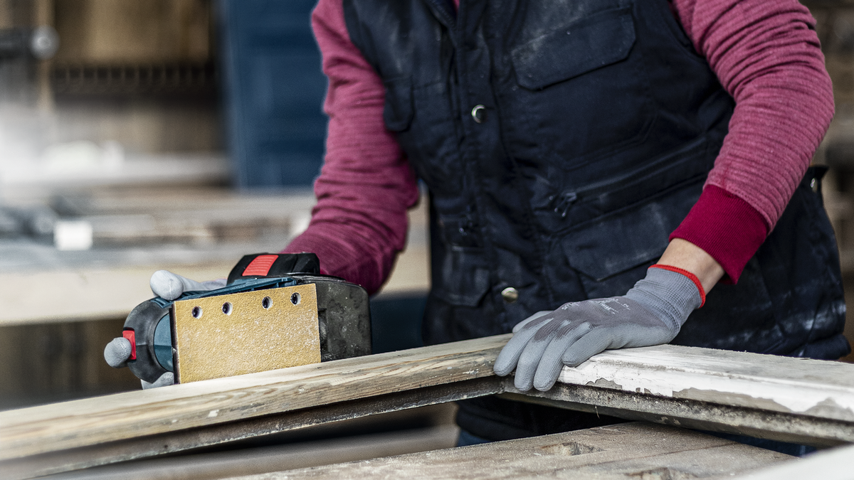 一名佩戴安全设备的人员正在用电动砂光机打磨木板。