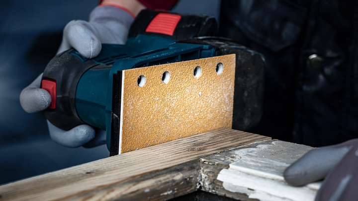 一名佩戴安全设备的人员使用手持式电动砂光机打磨木材。