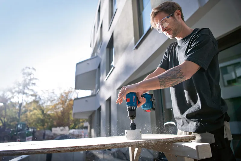 一名佩戴安全设备的人员在户外使用电钻在木头上钻孔。.