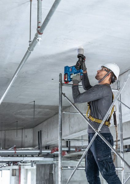 一名佩戴安全设备的人站在脚手架上，用电钻钻天花板。.