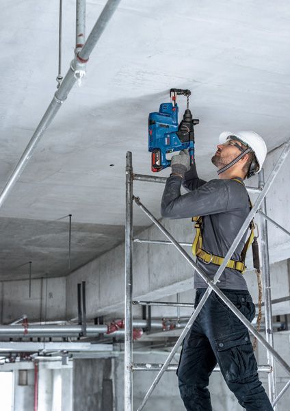 一名佩戴安全设备的人员站在脚手架上，向混凝土天花板上钻孔。.