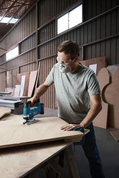 一名佩戴安全设备的人员在车间使用无绳曲线锯切割胶合板。.