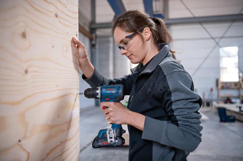 一名佩戴安全设备的工人在车间内钻木板。.