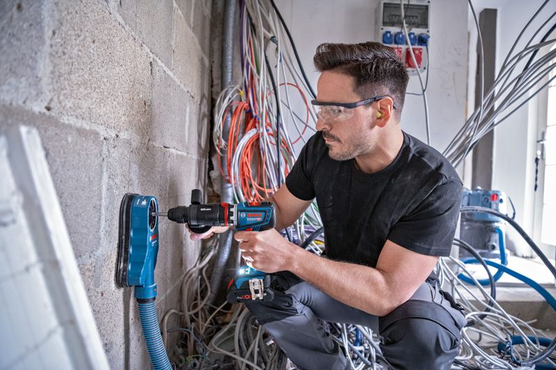 一名佩戴安全设备的人员正在用除尘器在混凝土墙上钻孔。.