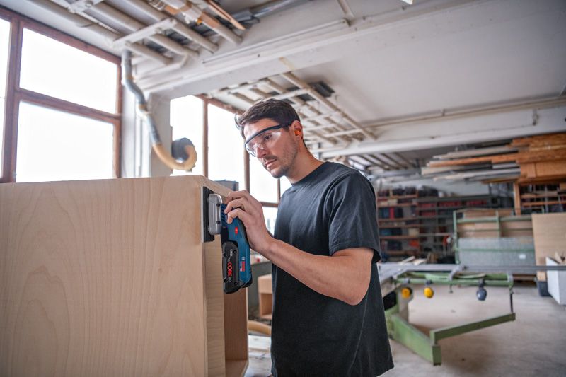 一名佩戴安全设备的人员正在使用无绳轨道砂光机打磨木制橱柜。.