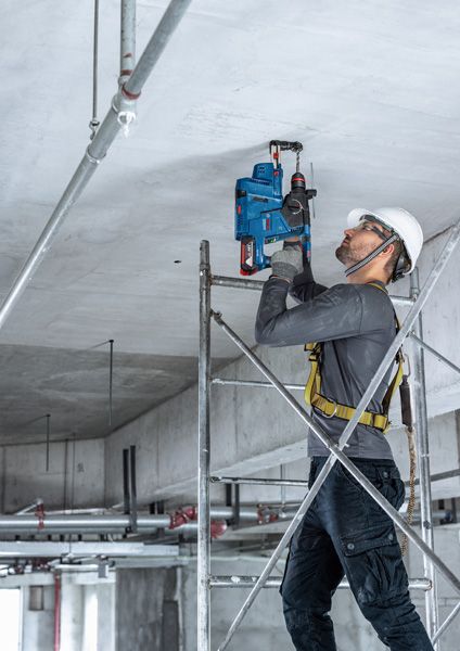 一名佩戴安全设备的人员站在脚手架上，向混凝土天花板上钻孔。.