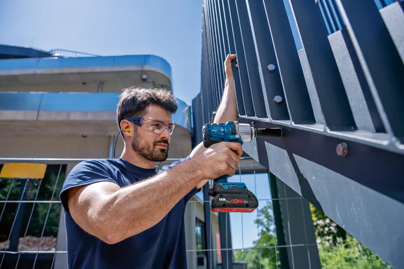 一名佩戴安全设备的人使用无绳冲击扳手撬动金属栅栏。.