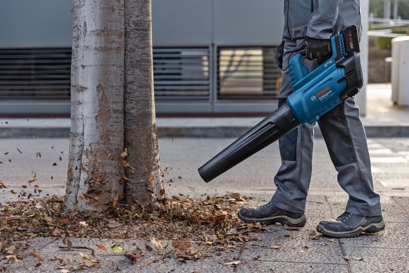 一名佩戴安全装备的人员使用无绳吹叶机清除树附近的落叶。