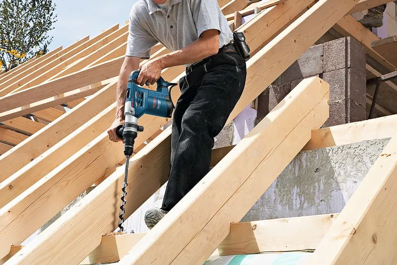 一名人员在屋顶结构的木梁上钻孔，没有佩戴安全设备。.