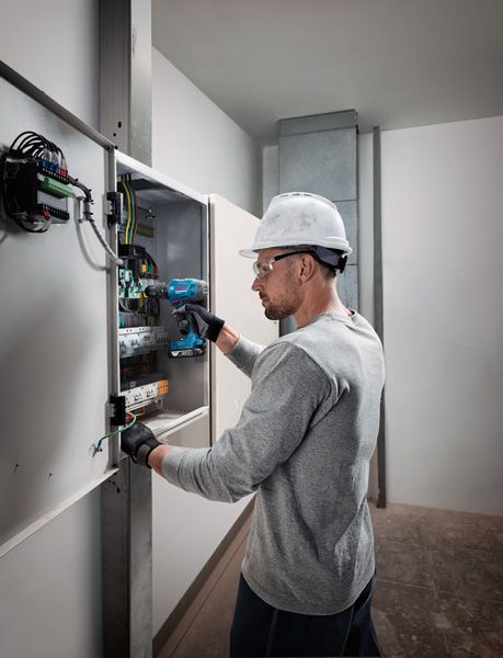 佩戴安全设备的人员使用无绳电钻在电气面板上工作。.