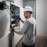 一名佩戴安全设备的人员使用无绳电钻在电气面板上工作。.