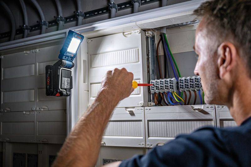 一个人使用螺丝刀在电气面板上操作，并连接无绳灯进行照明。.