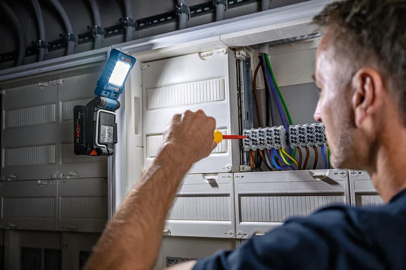 一个人使用螺丝刀在电气面板上操作，并连接无绳灯进行照明。.