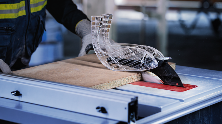 车间里，穿着安全设备的人员正在引导木材通过台锯。