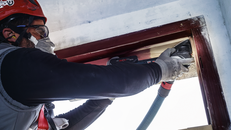 一名佩戴安全设备的人员正在使用电动工具打磨木制窗框。