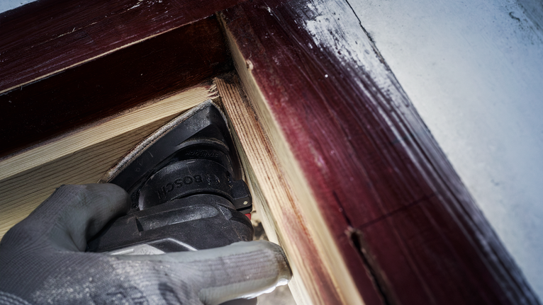 一名身穿安全设备的人员在油漆过的窗框附近打磨木材。