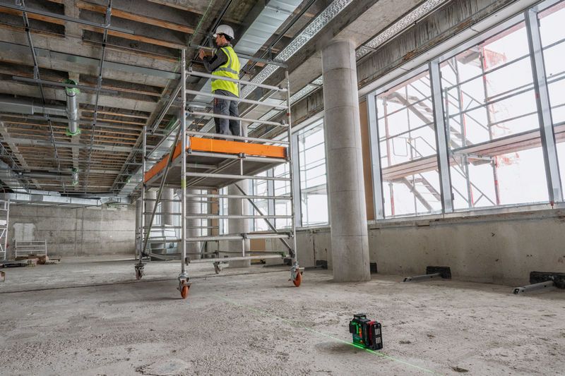 一名佩戴安全设备的人员在脚手架上工作，并使用激光调平工具投射线条。.