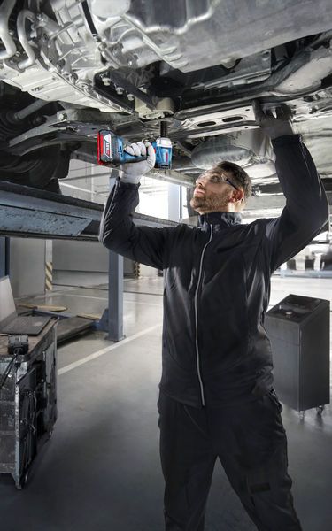 一名佩戴安全设备的人员使用电钻在升起的车辆下方作业。.