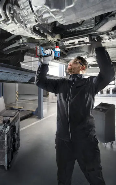 一名佩戴安全设备的人员在升起的车辆下方使用无绳电钻。.