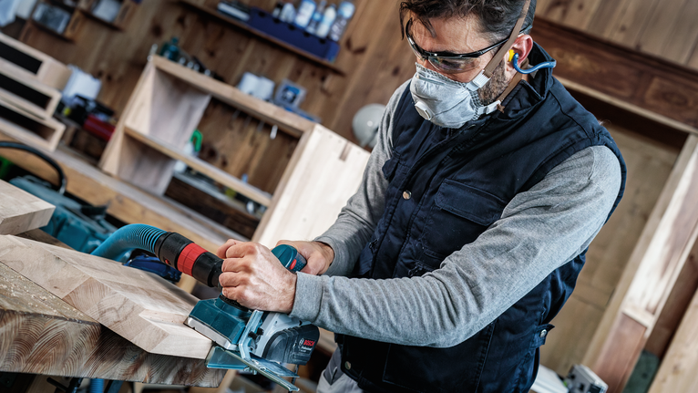 佩戴安全设备的人员使用电刨来打磨木板。