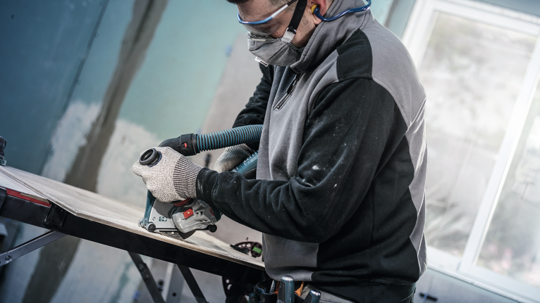 佩戴安全设备的人员使用连接到真空软管的电动工具修剪木材。