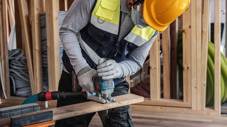 建筑工地上，一名佩戴安全设备的人员正在使用电动工具切割木材。