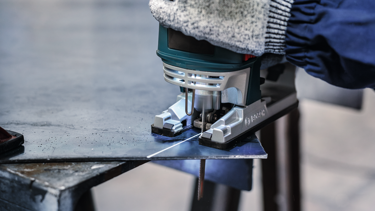 一名佩戴安全设备的人正在用曲线锯切割一块金属板。