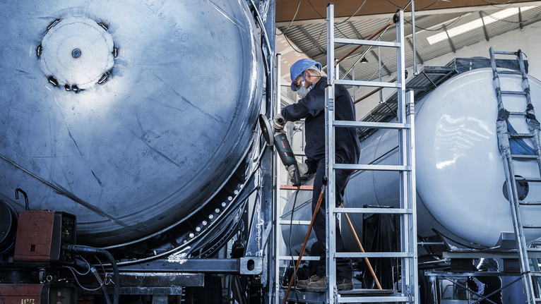 一名佩戴安全设备的人员使用电动工具对大型金属罐进行操作。