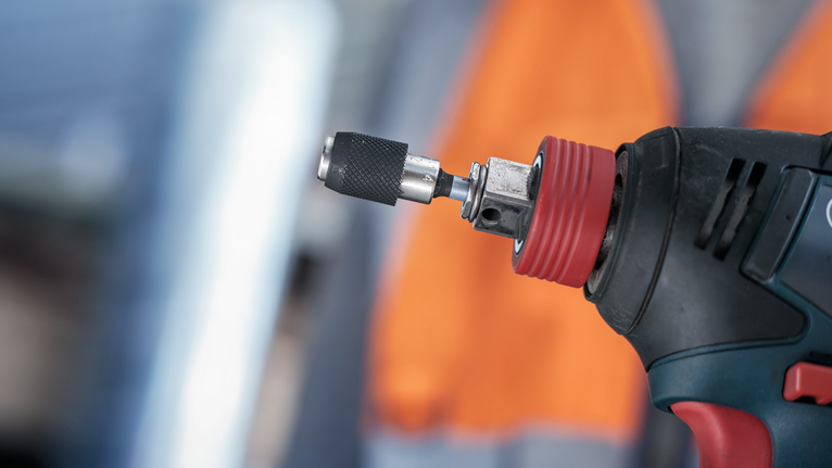 图中显示了带有金属钻头的电动工具，背景中的人佩戴着安全设备。